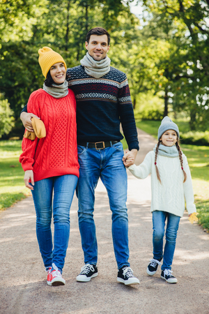 Portrait of friendly family walk across park. Beautiful brunette female in knitted yellow hat and sweater, handsome man embraces his wife and holds daughter`s hand, enjoy autumn weather and fresh airの写真素材