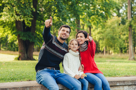Father and mother sit between their daughter against trees or nature background, show something to her, indicate with fore fingers. Smiling parents show bird to little beautiful girl. Parenthoodの写真素材