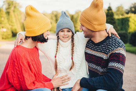 Adorable little girl with two pigtails, wears warm knitted hat and sweater, embrace her affectionate parents, admire beautiful nature, spend weekends together. People, happiness, relationshipの写真素材