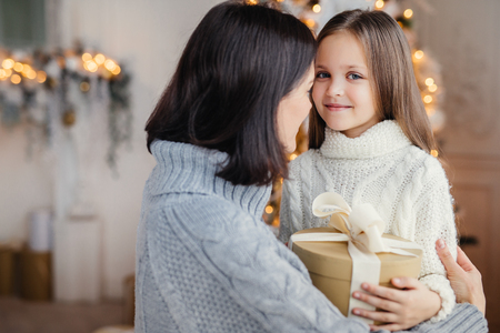 Young female embraces her little child, gives present, congratulates with New Year, proud to have such beautiful and clever daughter. Happy kid spends time with mother. Childhood and holidaysの写真素材