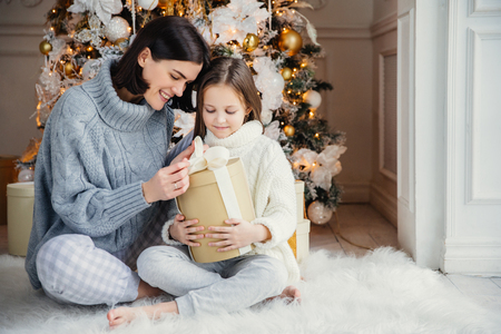 Indoor shot of adorable girl and her mother sit crossed legs, open wrapped present box, have intrigue what is there, sit against adorned New Year or Christmas tree, have sincere gentle smiles on facesの写真素材