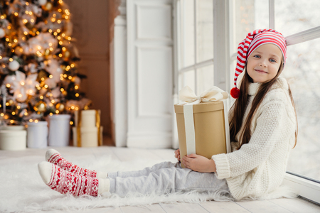 Cute lovely girl with present, sits near window in living room, looks delightfully into camera, admires wonderful beautiful decorated fir tree, has good mood and holidays. New Year conceptの写真素材