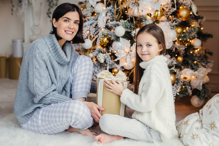 Little child and her mother sit on warm white carpet near decorated New Year tree, holds present in hands, have pleased and happy expressions, glad to spend holidays together. Childhood and holidaysの写真素材