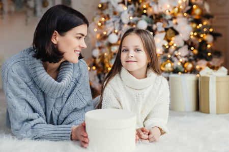 Winter and celebration concept. Pretty lovely blue eyed small female child in knitted white sweater and brunette female have good rest at home, spend time near adorned Christmas treeの写真素材