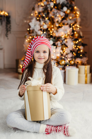 Vertical portrait of beautiful pleasant looking small child wears knitted sweater and socks, sits crossed legs with present, has desire to wrap it, being in living room near decorated New Year treeの写真素材