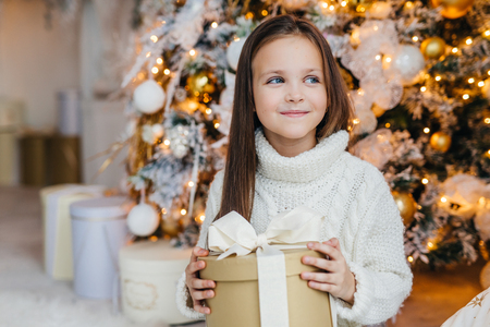 Cute lovely small kid with charming appearance, glad to recieve Christmas gift, looks aside with happy expression, has intrgue what present is, poses near beautiful decorated fir tree.の写真素材
