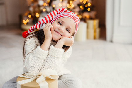 Beautiful small child poses in living room, leans at present gift, has happy expression, glad to recieve surprise from parents, spends holidays in family circle. Merry Christmas and happy New Yearの写真素材