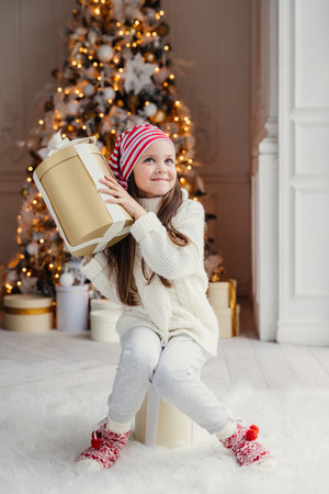 Curious small child doesn`t know what is in present box, recieves gift from Santa Claus, wears warm knitted clothes, sits against decorated New Year tree background. Childhood, surprisment, holidaysの写真素材