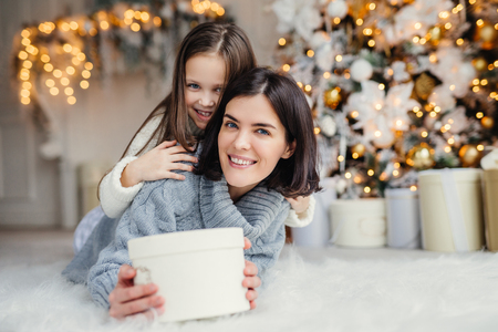 This present is for you! Happy small kid embraces her affectionate mother who holds wrapped present, stands against decorated background with garlands and New Year tree. Domestic atmosphere.の写真素材