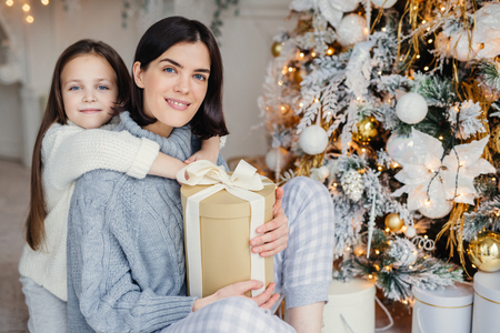 Thankful small female child embraces her mother who gave present, spend wonderful unforgettable time together, celebrate Christmas. Brunette woman and daughter look for present gifts under fir treeの写真素材