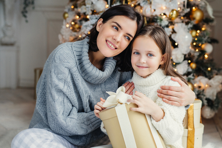 Cheerful brunette female leans at her daugter, embraces her, presents gift box, being in living room near decorated New Year tree. Glad family: mother and daughter in warm sweaters celebrate Christmasの写真素材