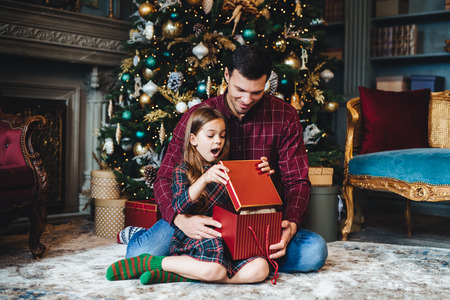 Surprised adorable small girl looks into present box, doesn`t believe her eyes, recieves pleasant gift from father, sit together on floor in cozy room near decorated New Year tree. Majestic momemtの写真素材