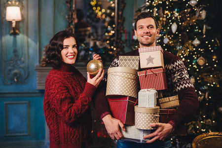 Smiling woman wears red knitted sweater holds glass ball as decorates fir tree, stands near her husband who holds piles of presents, going to present them to friends and relatives. Busy coupleの写真素材
