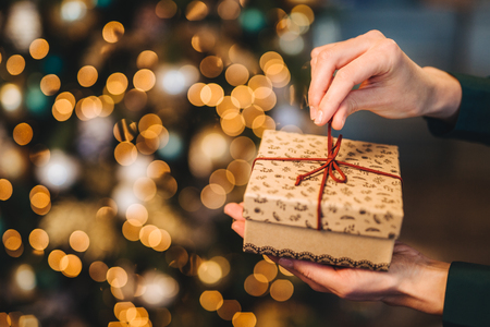 Unrecognizable woman wrappes gift box over Christmas tree with shining lights and garlands. Decorated present box in woman`a hands. Holidays and celebration concept.の写真素材