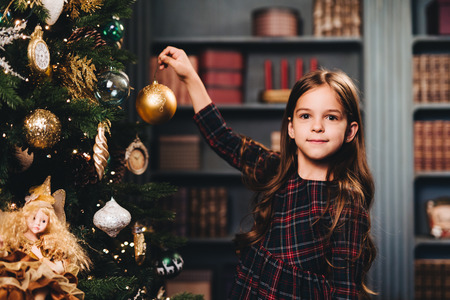 Small little kid decorates Christmas tree indoors, looks directly into camera, holds glass ball, being glad to have winter holidays. Little adorable girl wears dress decorates New Year tree aloneの写真素材
