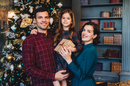 Happy family stand together against decorated fir tree, look happily into camera, hold present, celebrate New Year in family circle. Small girl being glad to spend holidays with her mother and fatherの写真素材