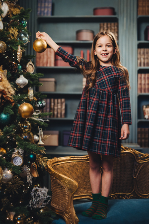 Smiling beautiful little girl stands on armchair, holds glass ball, decorates New Year tree, being happy to do it. Adorable small child busy with decoration of Christmas tree. Season and celebrationの写真素材