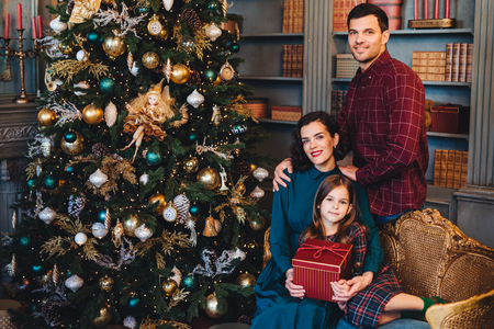 Beautiful brunette female embraces her little daughter and her husband sit together near decorated New Year tree, being glad to spend holidays in family circle. Affectionate family with happy looksの写真素材
