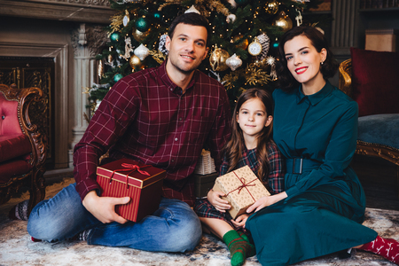 Father, mother and daughter enjoy holidays together, smile happily as sit in living room near decorated New Year tree, hold present wrapped in boxes, look delightfully into camera. Family conceptの写真素材