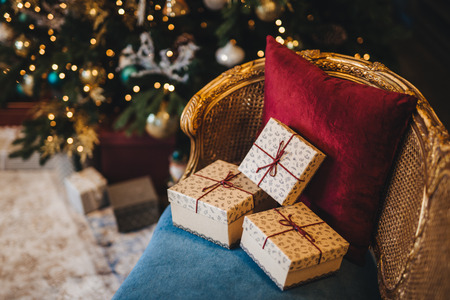 Three wrapped present boxes on armchair near Christmas fir tree. Design or idea for postcard. New Year composition with decorated fir tree and gifts. Majestic atmosphere. Festive eventの写真素材