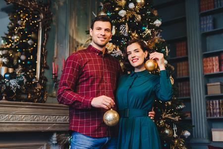 Handsome man with stubble embraces his beautiful wife, hold glass balls in hands, decorate New Year tree together, prepare for holidays. Smiling young beautiful female and her husband at homeの写真素材