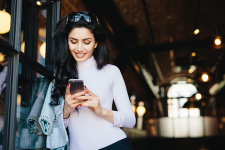Pretty Caucasian woman with dark wavy hair wearing sunglasses and white elegant clothes holding smartphone using wireless Internet connection while chatting with her friends. Youth, lifestyle conceptの写真素材