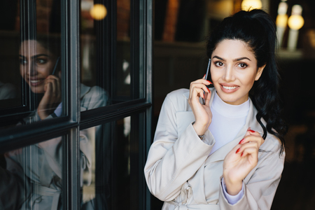 Pretty female with black hair having thin dark eyebrows, bright eyes with long eyelashes and full well-shaped lips wearing elegant clothing holding smartphone speaking with somebody and smilingの写真素材