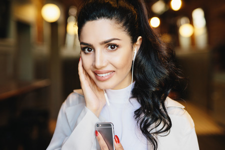 Close up portrait of attractive brunette woman with wavy pony tail, dark eyes and sensual lips having earphones in ears using app to connect with friends on social media. Lifestyle and fashion conceptの写真素材