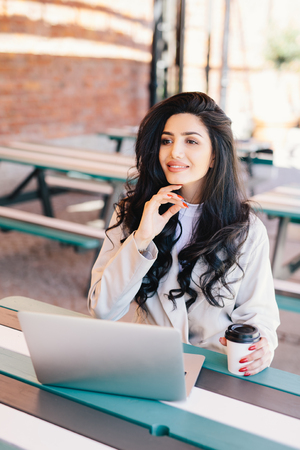 Elegant successful woman freelancer dressed formally using generic laptop computer for remote work, having rest at outdoor cafe drinking delicious coffee having dreamy expression. Lifestyle conceptの写真素材