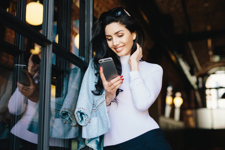 Young petty brunette businesswoman in elegant clothes standing against cafe interior waiting for people to have meeting using smart phone indoors, texting messages online. Communication conceptの写真素材