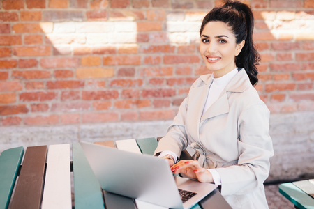 Pretty brunette student female wearing elegant white coat working at computer writing her thesis looking with smile at camera. Businesswoman in elegant clothes sitting indoors over brick wallの写真素材