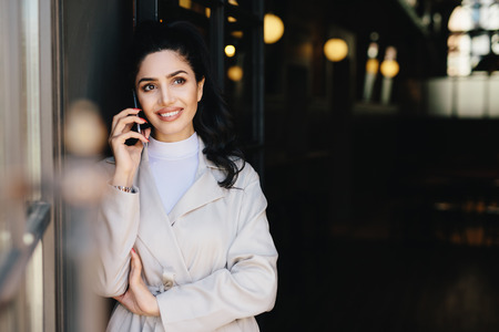 Elegant brunette businesswoman with dark eyes and full lips dressed in white clothes communicating over telephone having charming smile looking up with thoughtful expression. Technology conceptの写真素材