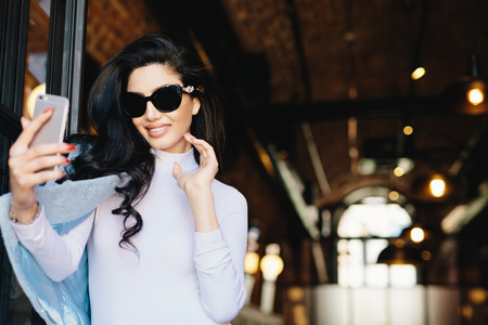 Smiling glamour woman in sunglasses, white blouse and jacket posing into camera of her smartphone while making selfie photographing herself. Happy elegant model posing in cafe using mobile phoneの写真素材