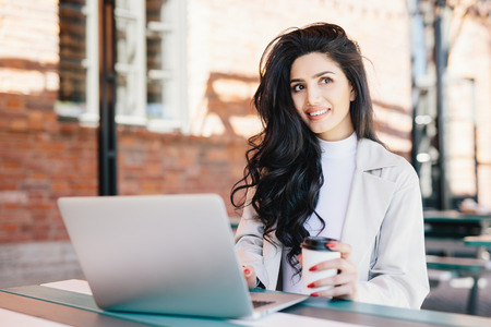Fashionable brunette woman with long luxurious hair dressed elegantly looking into distance with thoughtful expression dreaming of holidays while working on laptop at outdoor cafe. Technology conceptの写真素材