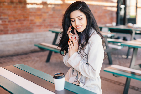 Lovely attractive woman with dark long hair, full lips and red manicure calling her friend with smartphone while having rest at cafe drinking coffee. Cute brunette female talking over cell phoneの写真素材