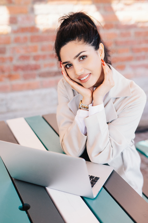 Indoor portrait of young successful Caucasian woman blogger writing a new post for her popular blog using notebook while sitting at desk over brick wall background. Smiling pretty lady with red nailsの写真素材