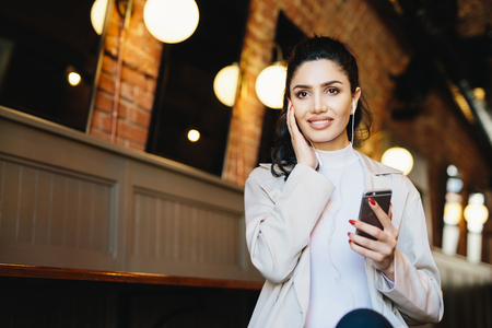 Graceful female with dark hair and eyes sitting in cafe using free internet conncetion while listening to music with earphones and mobile phone. Elegant lady using modern technologies indoorsの写真素材