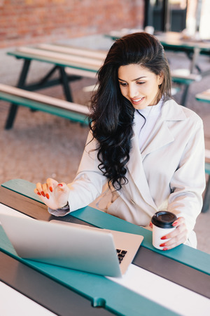 People and lifestyle concept. Top view of cute brunette female in white coat having red manicure sitting at table working with laptop computer, looking happily into screen drinking delicious coffeeの写真素材