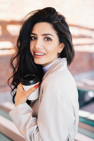 Vertical portrait of charming luxurious young lady with dark voluminous hair and appealing appearance holding takeaway coffee while waiting for her boyfriend going to have walk outdoors with himの写真素材