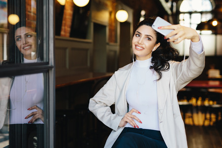 Waist up portrait of beautiful lady with dark hair tied in pony tail wearing white jacket sitting in cafe making selfie using her modern smartphone and listening to music with earphones. People, restの写真素材