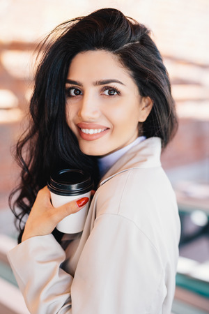 Cheerful female with dark hair lovely appearnce holding takeaway coffee wearing white coat. Smiling beautiful woman looking elegant and magnificent. People, lifestyle, relaxation, beauty conceptの写真素材