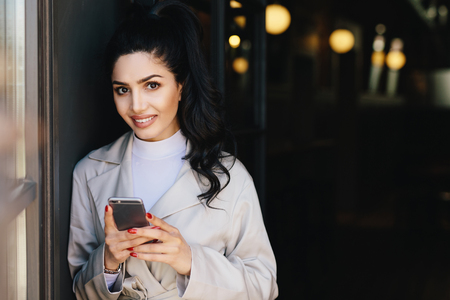 Gorgeous brunette woman with dark eyes, nice make-up and full lips dressed in white raincoat holding modern cell phone surfing social networks online. People, youth, lifestyle, technology conceptの写真素材