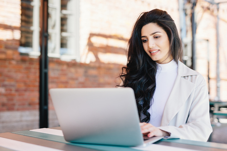 Adorable brunette woman in white clothes sitting outdoors at cafe using her laptop computer communicating with friends online using free internet connection. People, lifestyle, technology conceptの写真素材