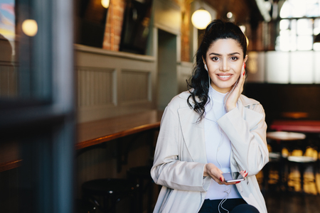 Happy brunette woman with make-up and manicure wearing white jacket holding smartphone and having earphones in ears listening to music or audiobook on earphones. Dreaming female in cafe using mobileの写真素材