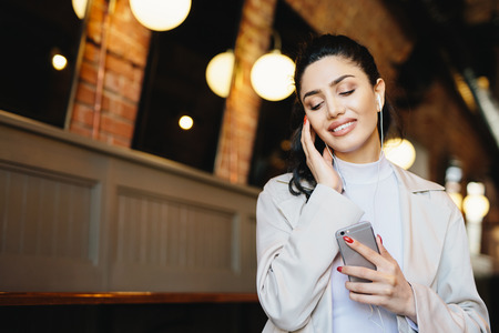 Portrait of beautiful female with dark hair tied in pony tail dressed formally listening to music with earphones closing her eyes with relaxation holding cell phone. People, lifestyle, technologyの写真素材