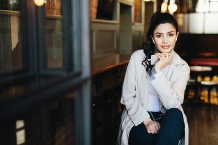 Attractive woman with dark hair and red manicure wearing stylish white clothes sitting in cozy cafe enjoying good tracks using free music app on her smartphone. People, leisure and lifestyle conceptの写真素材
