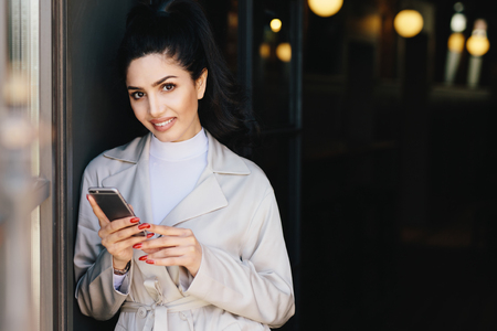 Portrait of adorable bruntte woman with dark eyes and eyebrows, pure healthy skin dressed in white clothes holding mobile phone in hands demonstrating her perfect manicure looking into cameraの写真素材