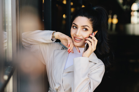 Attractive female with dark hair and nice appealing eyes having full lips dressed in elegant clothes having conversation with her best friend over cell phone looking aside. People, beauty conceptの写真素材