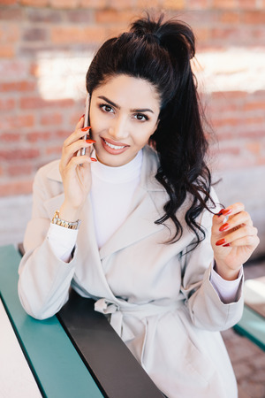 Young charismatic woman with dark wavy pony tail having red manicure dressed eleganltly talking on her mobile phone with smile while sitting at cafe waiting for her order. Communication conceptの写真素材