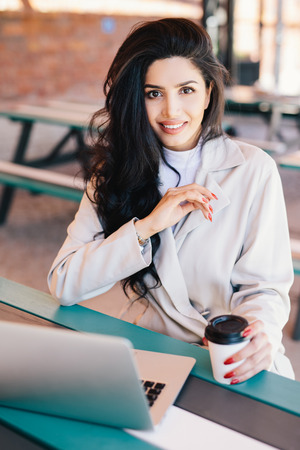 Vertical portrait of young brunette female entrepreneur enjoying rest at cafe with electronic device and coffee checking email on laptop, using free wireless connection looking directly into cameraの写真素材